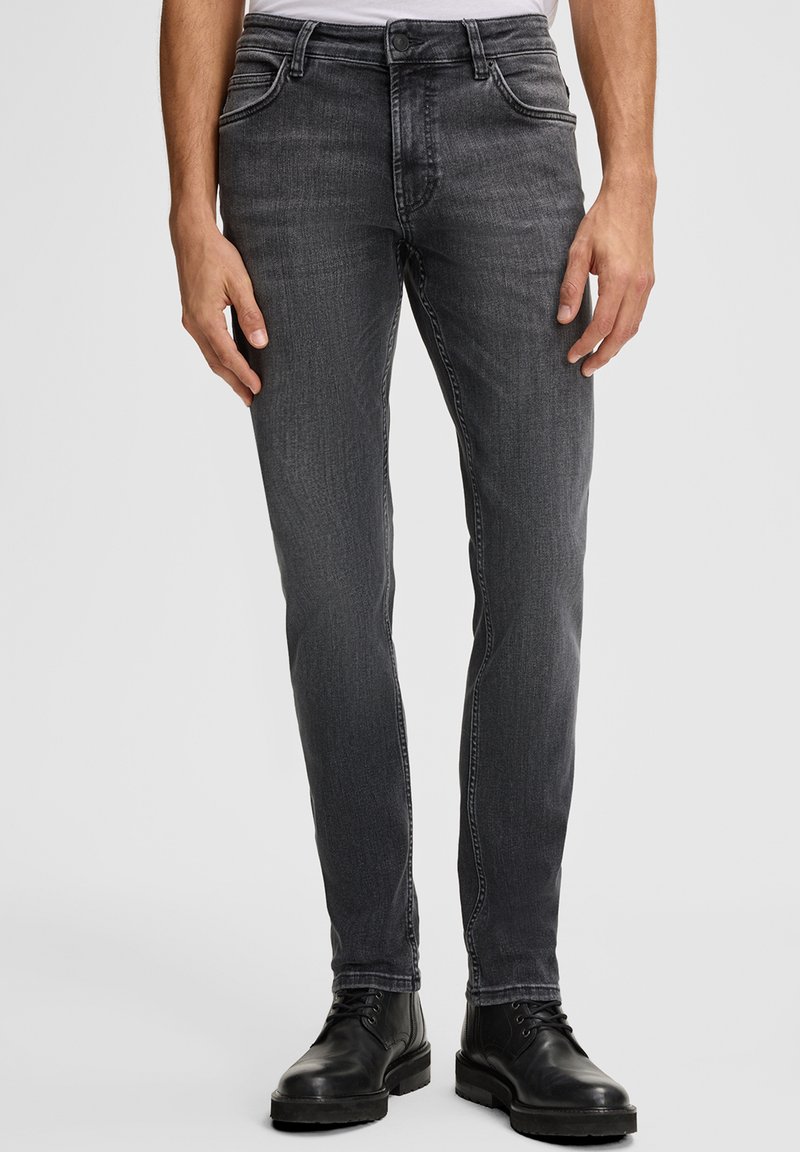 Uomo che indossa jeans neri slim-fit sbiaditi e stivali in pelle neri con lacci, in piedi contro uno sfondo grigio chiaro semplice.