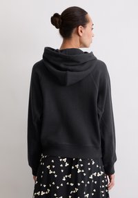 Felpa nera con cappuccio, dal taglio morbido, con polsini e orlo a coste. Tessuto liscio, indossata sopra una gonna a fiori.