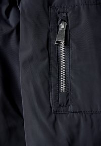 Zbliżenie na częściowo otzipowany srebrny zamek błyskawiczny w kieszeni czarnej tkaninowej kurtki z wyraźnym przeszyciem.