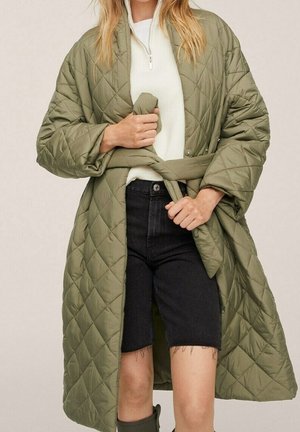 Kvinde binder bælte på lang olivengrøn quiltet frakke, iført hvid sweater og sorte knælange denimshorts.