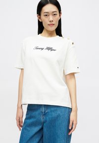 Λευκό βαμβακερό t-shirt με κοντά μανίκια, με μαύρη κεντητή επιγραφή "Tommy Hilfiger" και χρυσές κουμπίδες στους ώμους.
