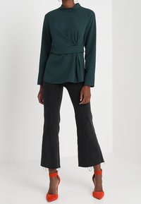 Tmavě zelený top s dlouhými rukávy a vysokým límcem s předním detailem s uzlem, kombinovaný s čern�ými rozšířenými džíny a jasně červenými botami na vysokém podpatku.