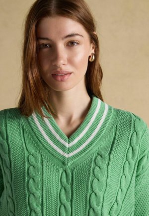 Young woman with light brown hair wearing a green cable knit sweater with white-striped V-neck and gold hoop earrings against beige background.