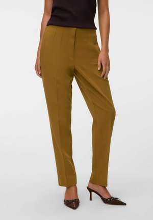 Femme portant un pantalon jaune moutarde sur mesure et un débardeur noir, avec des chaussures marron à talons et bouts pointus.