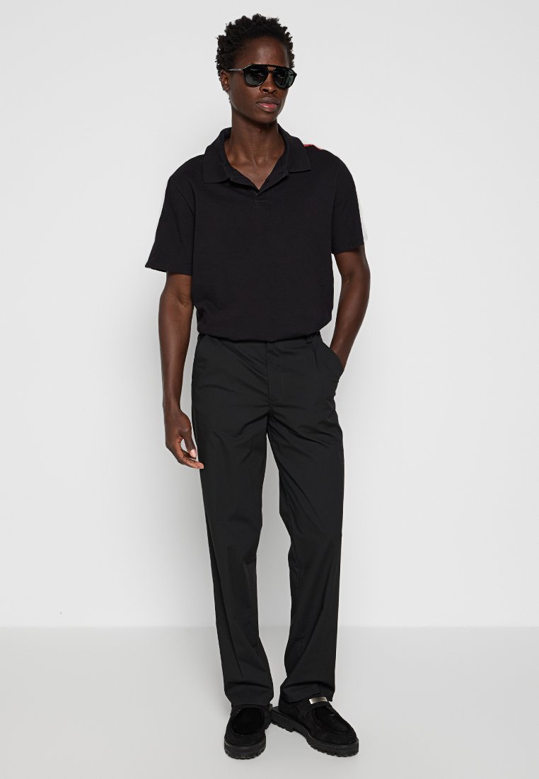 Chemise polo noire avec un col, associée à un pantalon noir taille haute et des chaussures noires. Lunettes de soleil portées ; accessoires minimaux.