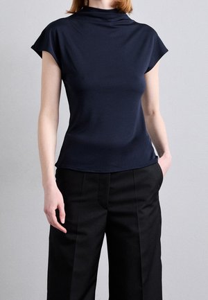 Vrouw draagt een marineblauw topje met korte mouwen en opstaande kraag en zwarte, hoog getailleerde, op maat gemaakte broek tegen een lichtgrijze achtergrond.