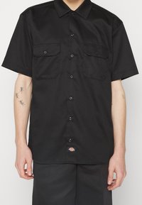 Chemise noire à manches courtes en tissu lisse avec deux poches poitrine, fermeture à boutons sur le devant et une étiquette sur l'ourlet inférieur.
