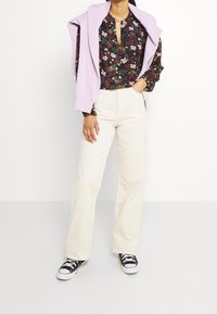 Blouse noire fleurie à encolure en trou de serrure, associée à un pantalon patte d'éléphant en velours c ôtelé écru et des baskets noires à semelles blanches. Pull violet clair posé sur les épaules.