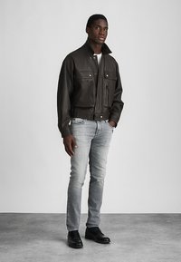 Καφετί δερμάτινο μπουφάν με κοντό γιακά, δύο τσέπες στο στήθος, μπροστινό φερμουάρ και κουμπί που κλείνει. Φορεμένο με γκρι τζιν και μαύρα παπούτσια.