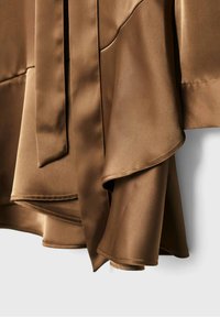 Tissu en satin marron avec des bords incurvés superposés et une finition lisse et brillante, mettant en valeur des coutures détaillées sur les manches du vêtement.