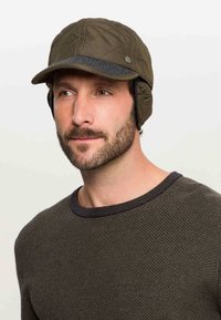 Man met een korte baard, die een gewatteerde olijfgroene wintermuts met oorkleppen en een gestructureerde donker olijfgroene trui draagt.