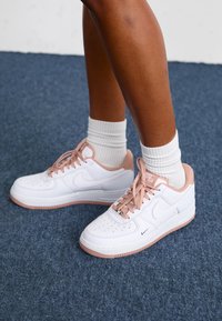 White Nike Air Force 1 sneakers with pink accents, perforated toe box, and brown rubber sole. Paired with white ribbed socks.