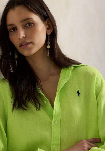 Γυναίκα με μακριά σκούρα μαλλιά που φοράει φωτεινό πράσινο πουκάμισο με κουμπιά και μακριά σκουλαρίκια με χάντρες, κοιτάζοντας ελαφρώς προς το πλάι πάνω σε απλό φόντο.