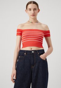 Top corto rosso scollato con strisce bianche; texture a costine. Abbinato a pantaloni in denim blu scuro a gamba larga e con una tasca.