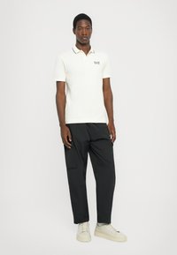 Crème polo shirt met zwarte details en het EA7-logo, gedragen met zwarte cargobroeken en lichtgrijze sneakers. Volledige lengte weergave van een mannelijke model.
