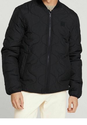 Mężczyzna w czarnej pikowanej kurtce na zamek z elastycznymi mankietami i jasnych spodniach, stojący z ramionami swobodnie opuszczonymi wzdłuż ciała.