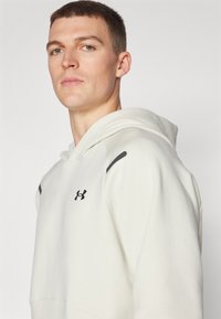 Weiße Kapuzenjacke aus weichem Material, mit einer Kängurutasche, rundem Ausschnitt und schwarzen Akzenten an den Schultern. Aufdruck des Under Armour Logos.