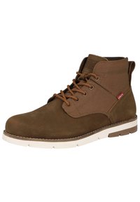 Bota de tobillo de cuero marrón y lona con puntera redonda, superficie texturizada, cordones marrones, suela de goma blanca y un acento de logo rojo.