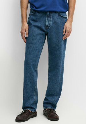 Mand iført blå jeans med lige ben, mørkebrune snørebåndssko og en blå skjorte, stående med armene afslappet ned langs siden.