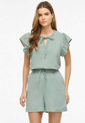 Žena v svetlozelenom textúrovanom top s krátkymi rukávmi s volánmi a zodpovedajúce vysokým pásom šortky viazané v páse.