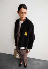 Zwarte wollen varsity-jack met witte strepen op de manchetten en onderrand. Bevat een gele geborduurde lintlogo. Neutraal-kleurig outfit daaronder.