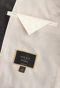 Interior de tela negra y crema de una chaqueta que presenta un bolsillo triangular y una etiqueta negra con texto dorado que dice "NEXT Occasion Slim Fit 52R".
