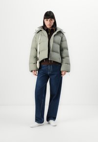Lys grøn pufferjakke med en cropped design og en aftagelig hætte, parret med en brun sweater og vide blå jeans. Hvide sko.