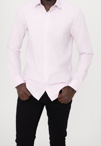 Camisa de botones a rayas en rosa claro con rayas blancas, con mangas largas y dobladillo curvado. Se lleva sobre pantalones negros.