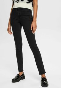 Jeans skinny negros de denim, con una cintura de altura media, líneas limpias y una silueta ajustada. Combinados con mocasines negros brillantes.