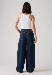 Jeans en denim bleu foncé à jambes larges et taille haute, avec une patch en cuir marron à l'arrière et deux poches arrière, portés avec un débardeur blanc.