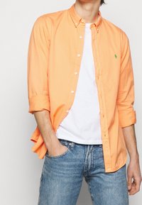 Oranžová košile na knoflíky s ohrnutými rukávy, lehký materiál, doplněná bílým tričkem a modrými denimovými džínami. Zelený logo akcent.