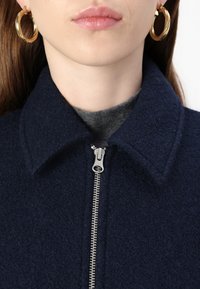 Cappotto di lana blu navy con colletto e zip argentata. Il modello indossa un top grigio sotto e orecchini a cerchio dorati. Tessuto morbido e texturizzato.