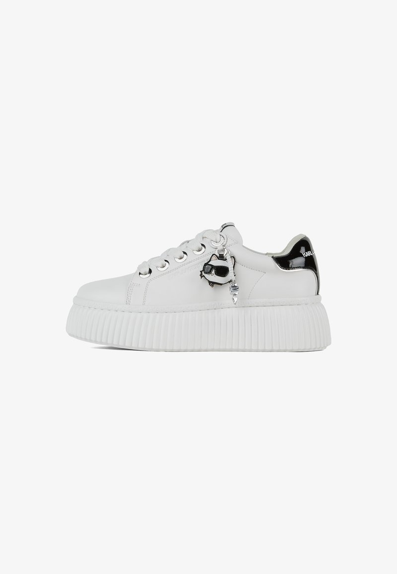 Weiße Sneakers mit einer strukturierten Plateau-Sohle, metallischen Ösen und einem schwarzen Lacklederspitzenelement. Enthält einen dekorativen Anhänger.