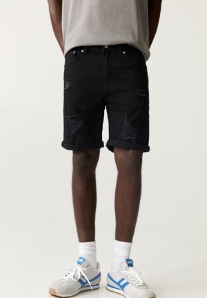 Schwarze aufgerollte, abgenutzte Jeansshorts, getragen von einer stehenden Person, kombiniert mit weißen Socken und weißen Turnschuhen mit blauen Streifen.