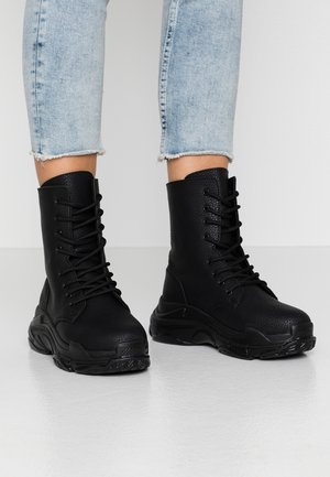 Schwarze, strukturierte Leder-Schnürstiefel im Combat-Stil, getragen zu hellblauen, gekürzten Jeans vor einem schlichten hellen Hintergrund.
