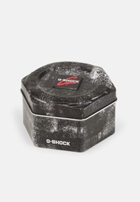 Recipiente hexagonal preto de estanho com uma superfície texturizada e detalhes em prata. Apresenta um logo vermelho da G-SHOCK na tampa.