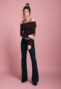 Bordeauxrotes, geripptes Off-Shoulder-Oberteil kombiniert mit dunklen ausgestellten Jeans, vor einem rosa Hintergrund. Das Modell trägt silbernen Schmuck.