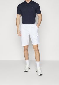 Marinblå polo med tre knappar och logotyp, i kombination med vita knälånga shorts och vita sportskor i lätt tyg med nätaccenter.