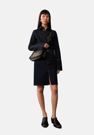 Abito camicia in denim scuro con spacco frontale, maniche lunghe e cuciture a contrasto, abbinato a una borsa a tracolla nera e zoccoli.