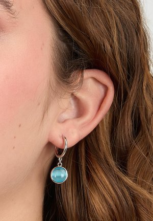 Primo piano di un orecchio con capelli castano chiaro che indossa un orecchino a cerchio in argento con un ciondolo rotondo in pietra azzurra chiara.