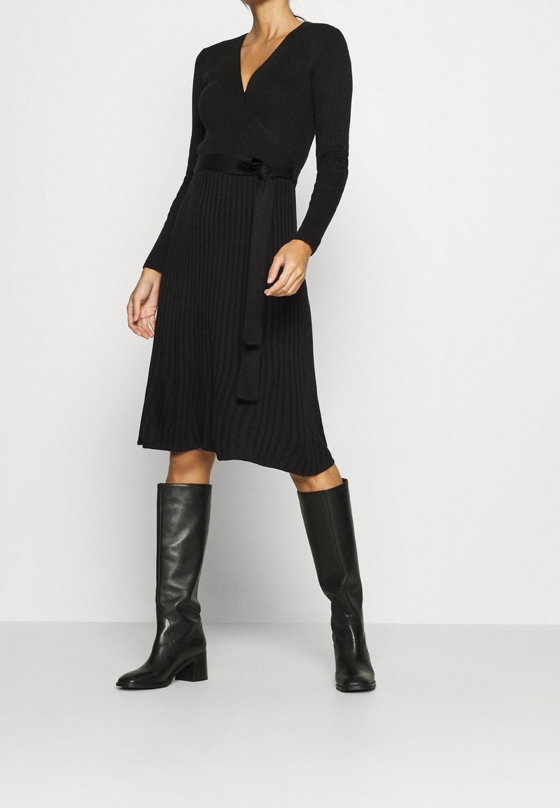 Femme portant une robe portefeuille en maille noire côtelée avec une ceinture et des bottes en cuir noires montant jusqu'aux genoux, debout devant un fond clair uni.
