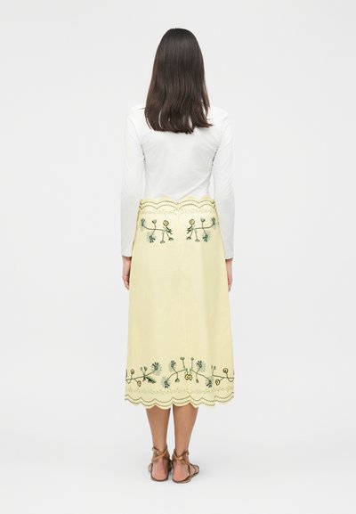 Γυναίκα με μακριά σκουρόχρωμα μαλλιά που φοράει λευκό μακρυμάνικο τοπ και παστέλ κίτρινη midi φούστα με κεντήματα λουλουδιών και κυματιστό τελείωμα, πλάγια όψη από πίσω.