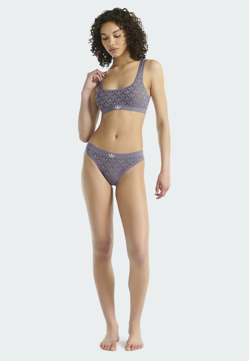 Žena s kudrnatými vlasy pózující bosá v odpovídajícím šedém sportovním podprsenkovém topu a kalhotkách s jemným potiskem trefoil Adidas na jednobarevném pozadí.