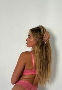 Set di lingerie in pizzo rosa composto da un reggiseno con spalline sottili e banda con logo, abbinato a un perizoma in pizzo coordinato, esposto su uno sfondo neutro.