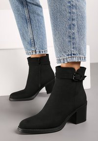 Bottines noires en daim, avec une fermeture éclair latérale, un accent de boucle, un bout carré et un talon épais. Portées avec un jean bleu.