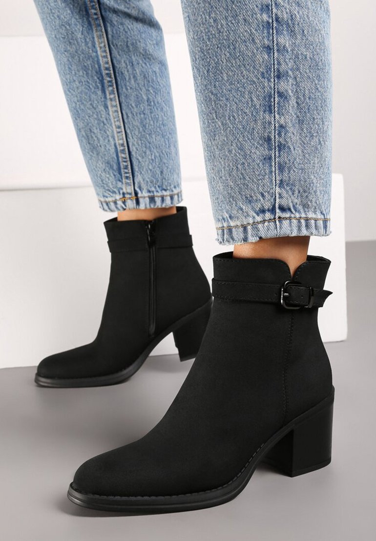 Bottines noires en daim, avec une fermeture éclair latérale, un accent de boucle, un bout carré et un talon épais. Portées avec un jean bleu.