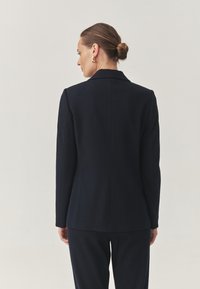 Námořnický blazer na míru se strukturovaným designem, klopovým límcem a dlouhými rukávy. Hladká textura a minimální švy pro elegantní vzhled.