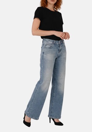 Donna che indossa un top nero a maniche corte, jeans azzurri a gamba larga e scarpe nere con tacchi alti, in piedi su uno sfondo semplice.