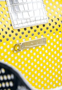 Geel geperforeerde tas met een zilveren nepkrokodillenaccent, metalen hardware en een geprinte logotag. Textuur, lichtgewicht ontwerp.