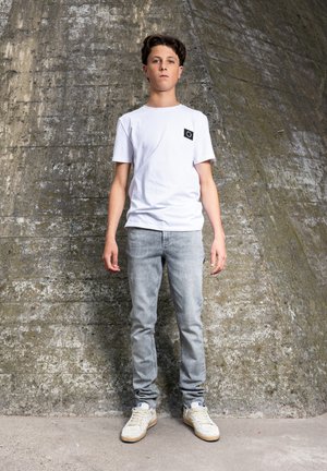 Hvid bomuld T-shirt med en sort lomme, lysegrå denimjeans og beige sneakers. Står op af en tekstureret betonvæg.